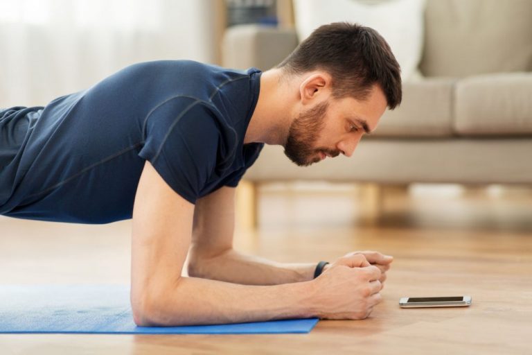 Mann macht Plank zuhause und schaut dabei auf sein Handy
